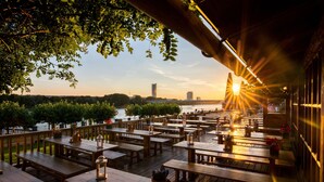 An outdoor dining area with wooden tables and benches, overlooking a body of water and a city skyline during sunset. - Kameha Grand Bonn (Bonn)
