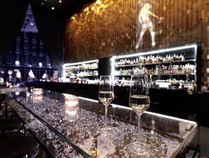 Un bar con dos copas de vino blanco sobre una superficie reflectante, una barra y estantes llenos de botellas al fondo.