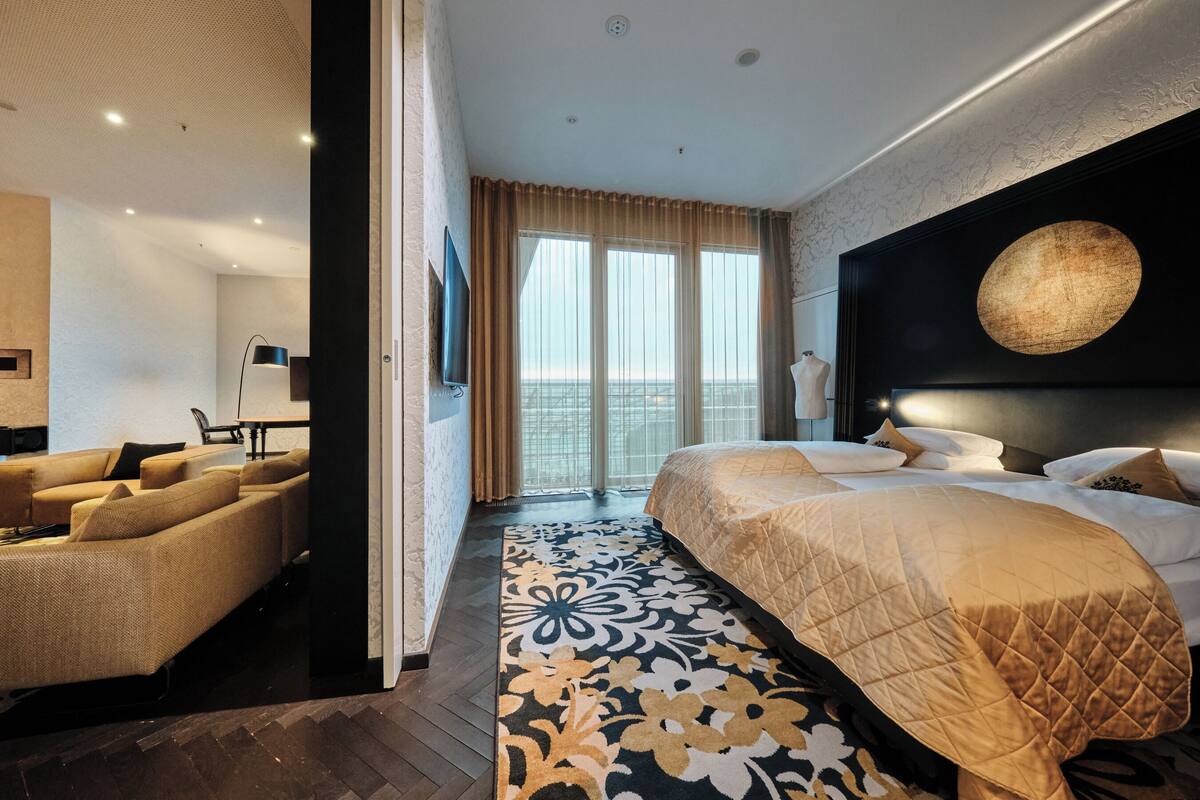 A modern hotel room with a large bed, a sofa, a dining table, and a view of the city.