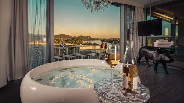 A room with a large window, a jacuzzi, a bottle of wine, and a television. - Kameha Grand Bonn (Bonn)