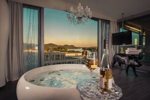 A room with a large window, a jacuzzi, a bottle of wine, and a television.
