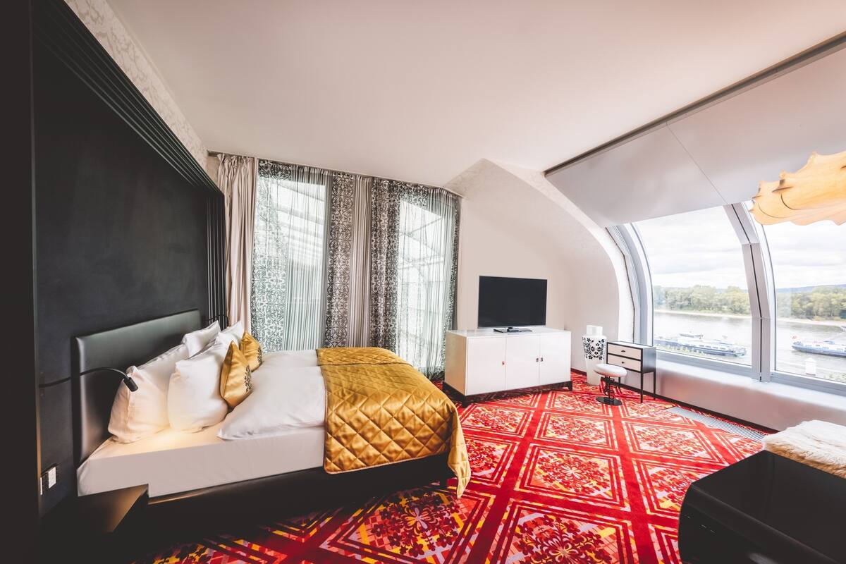 A modern hotel room with a large bed, a TV, a desk, and a view of a river.