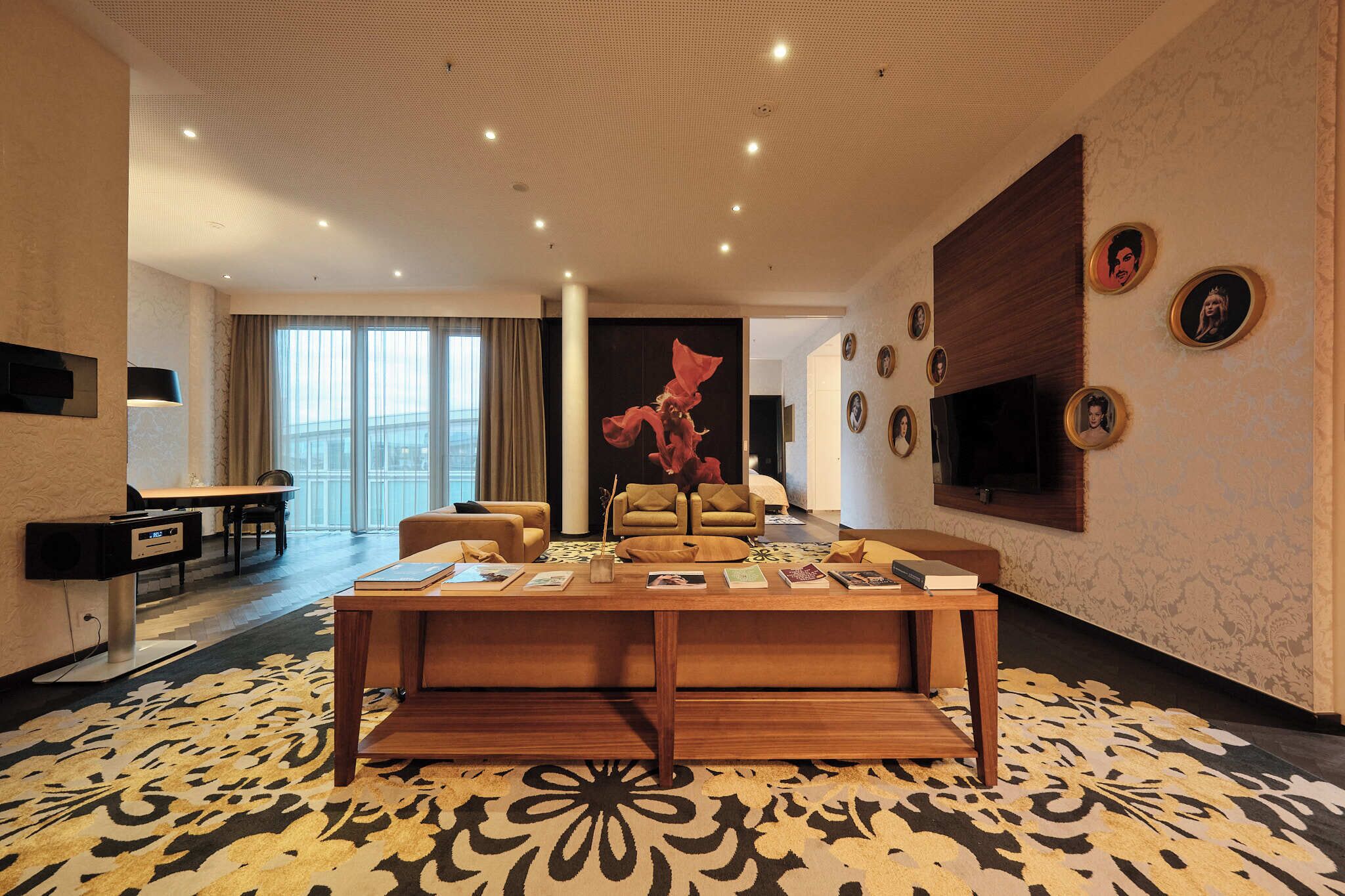 Una habitación de hotel moderna con un ventanal grande, un televisor de pantalla plana montado en la pared, una mesa de centro de madera con libros y una alfombra estampada.