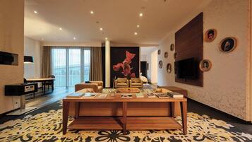 A modern hotel room with a large window, a flat-screen TV mounted on the wall, a wooden coffee table with books, and a patterned carpet.
