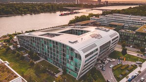 A modern building with a curved facade and large windows, situated near a river with a bridge and boats in the background. - Kameha Grand Bonn (Bonn)