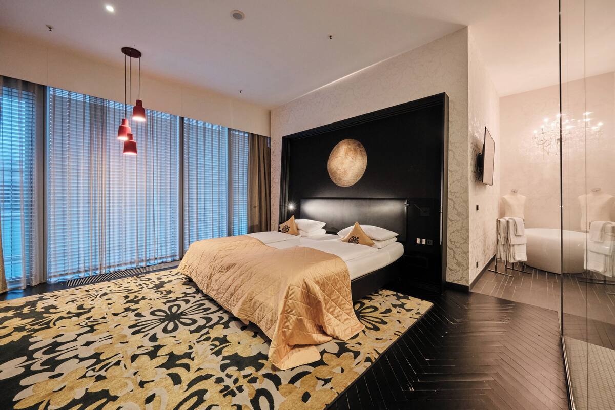 A modern hotel bedroom with a large bed, a patterned rug, and a view of the city through large windows.