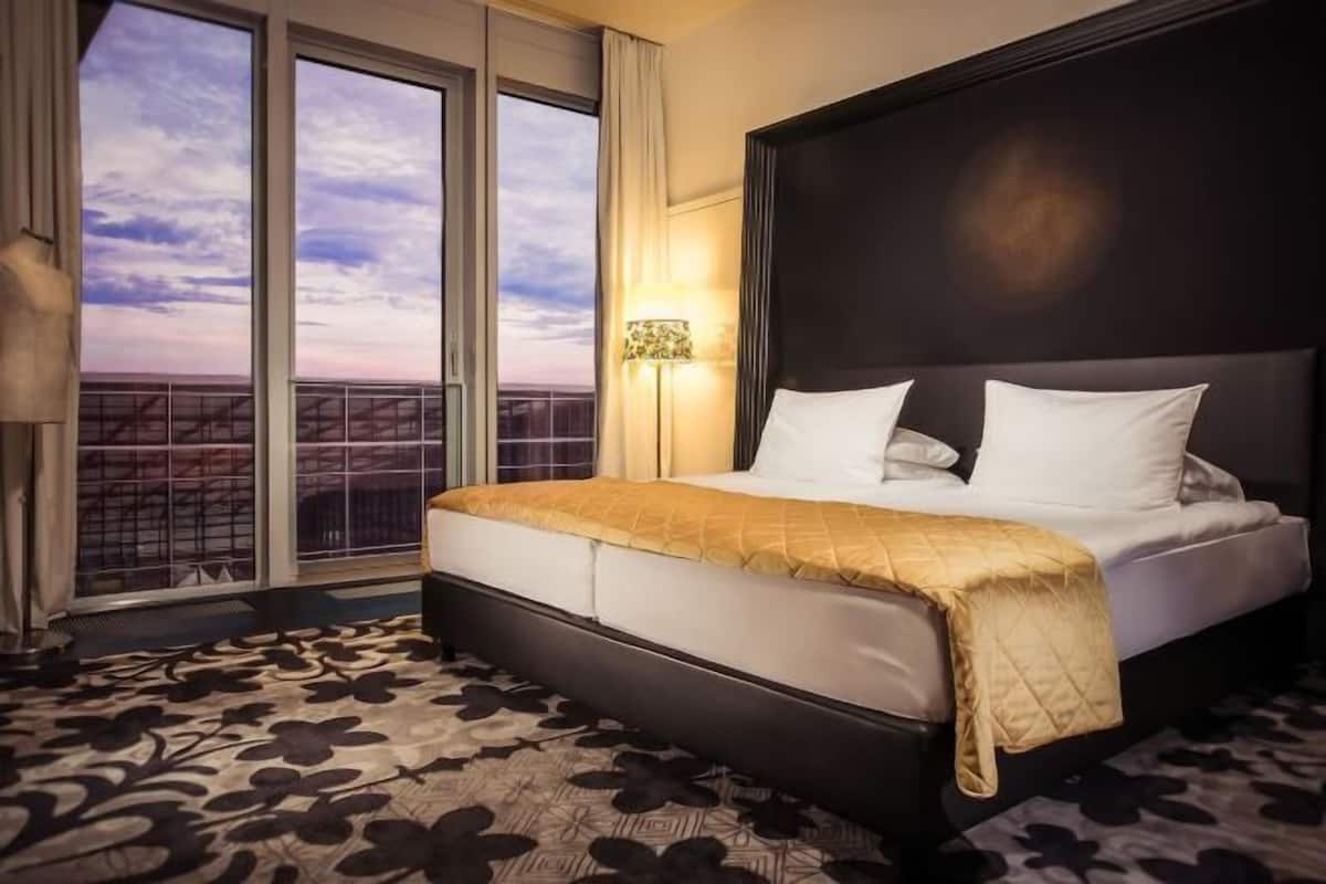 A hotel room with a large bed, a view of the cityscape through floor-to-ceiling windows, and a patterned carpet.
