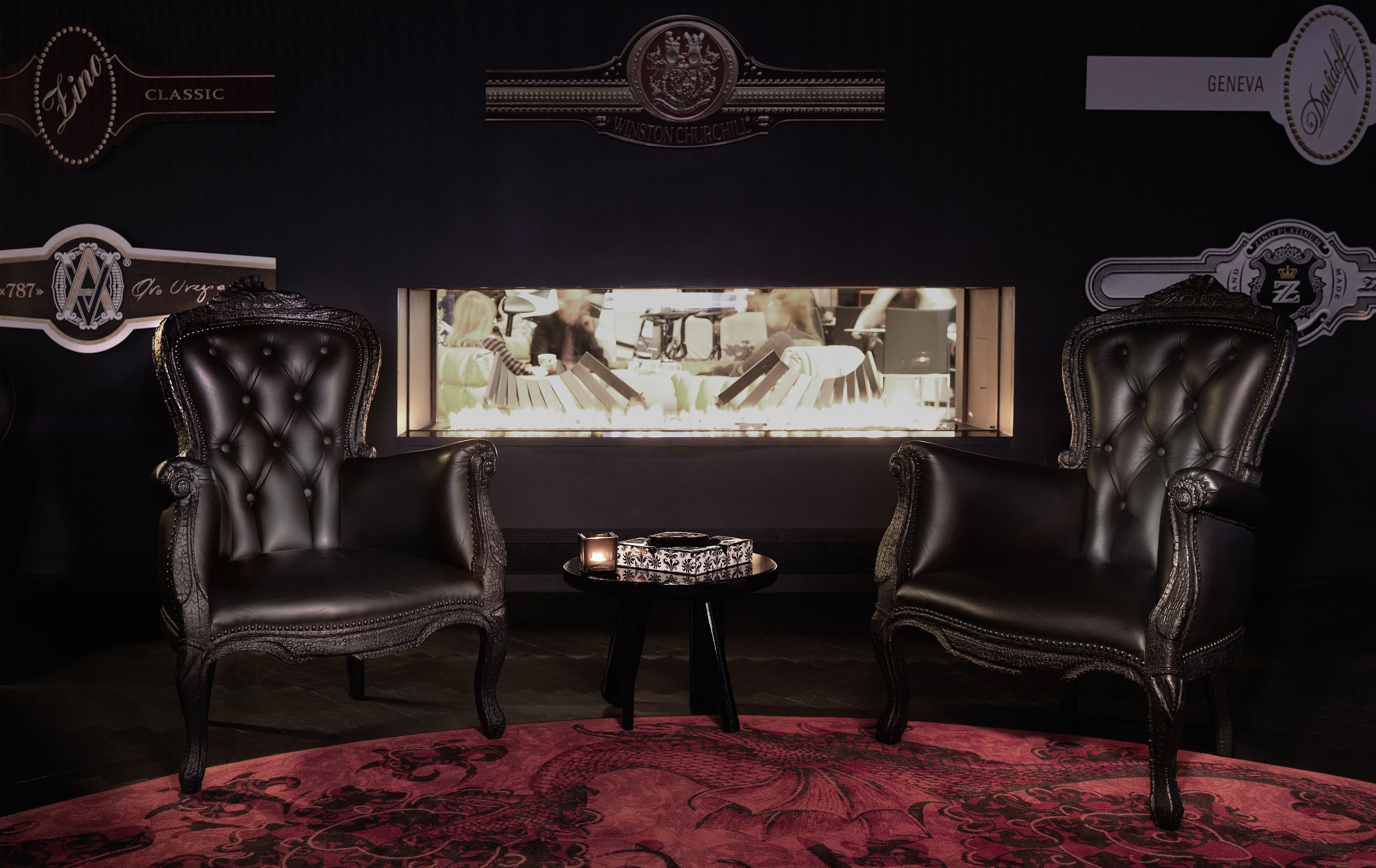 A dark room with two ornate leather chairs, a small table with a patterned tablecloth, and a television screen displaying a scene.