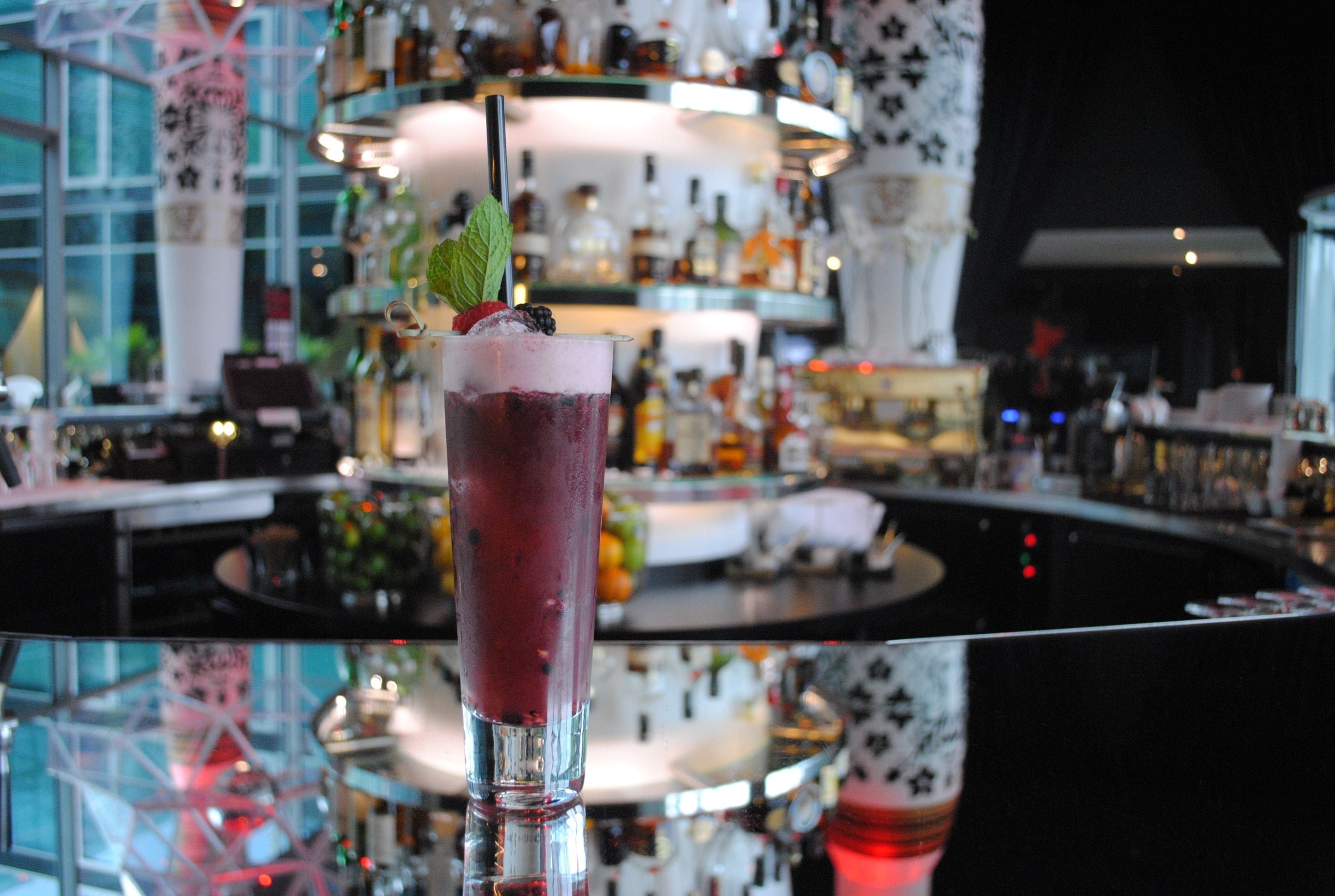 Un bar con un bicchiere alto contenente una bevanda viola guarnita con una foglia di menta e una fragola, sullo sfondo scaffali pieni di diverse bottiglie.