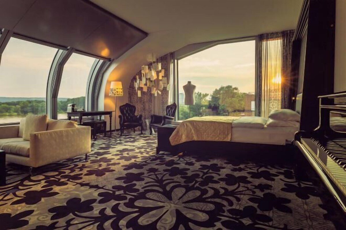 A hotel room with a large bed, a piano, a desk, and a view of the outdoors.