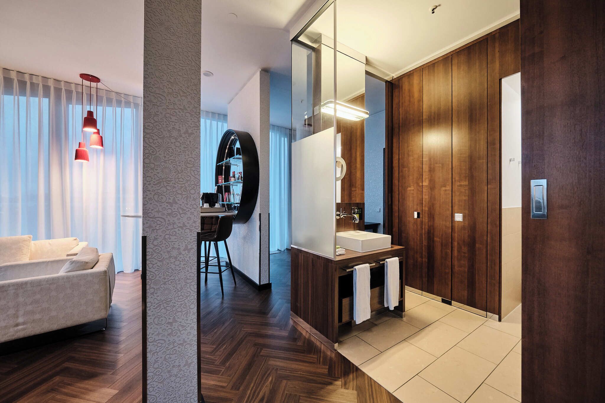 A modern hotel room with a sofa, a dining area, a large mirror, and a wooden wardrobe.