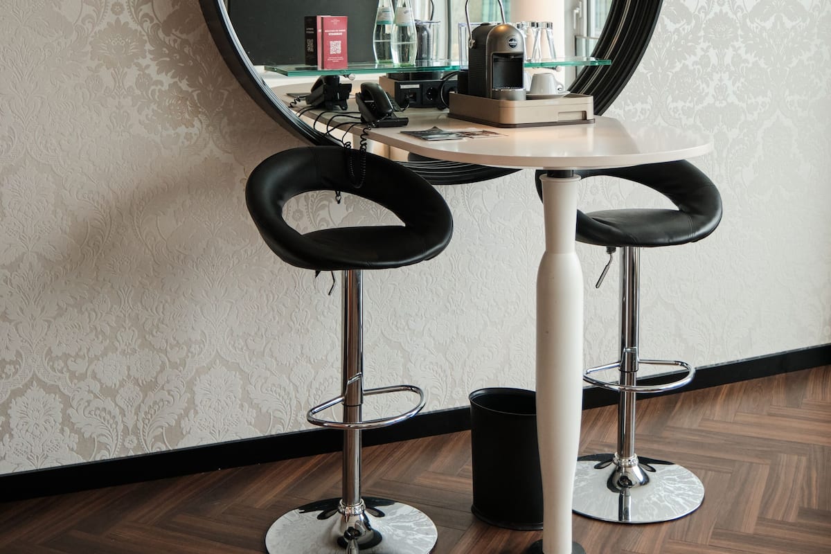A room with a round mirror, two black bar stools, a white table, and a patterned wallpaper.