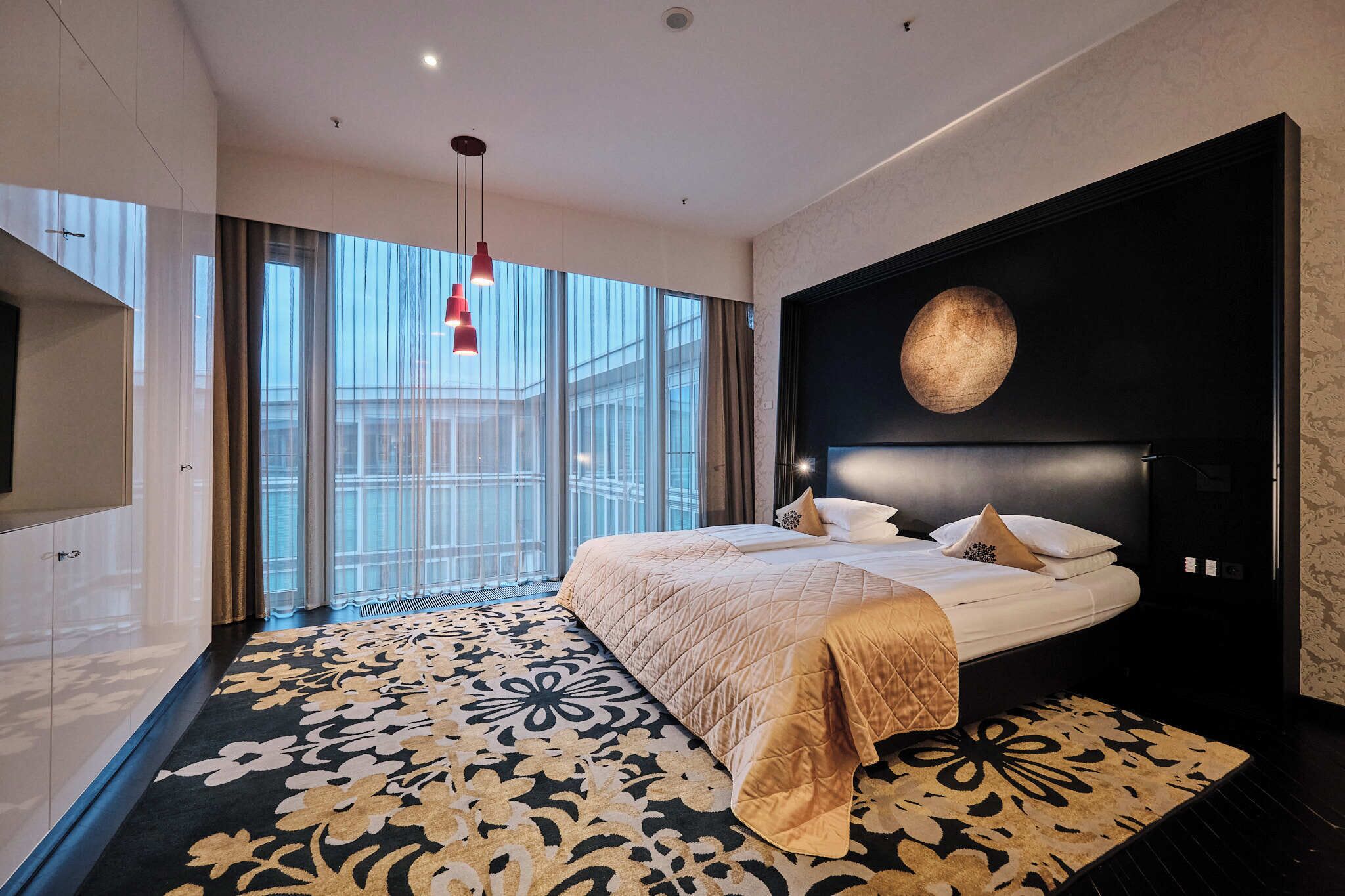 A modern hotel room with a large bed, a TV, and a view of the city through large windows.