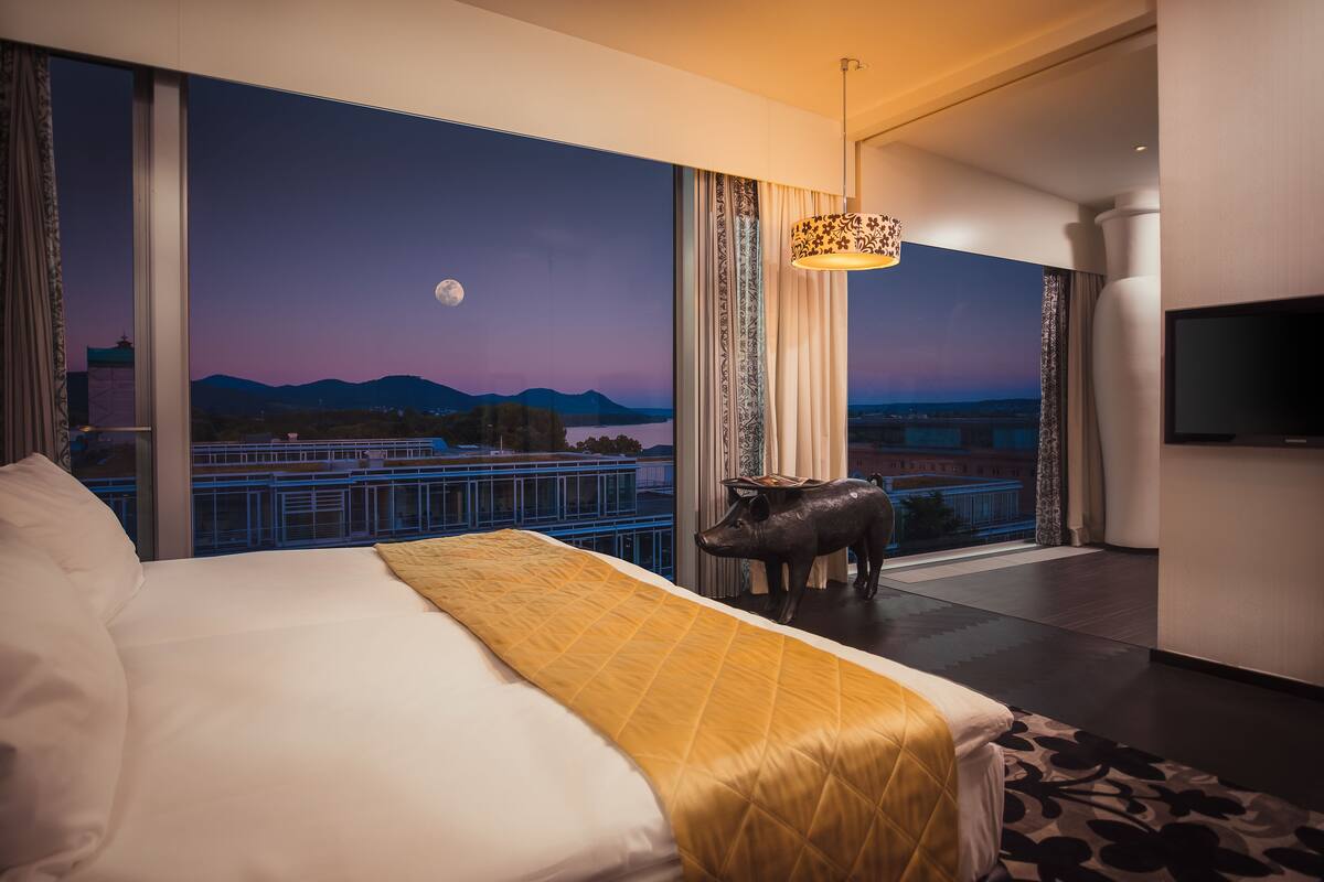 A hotel room with a large bed, a view of the cityscape at dusk, a mounted pig sculpture, and a television on the wall.