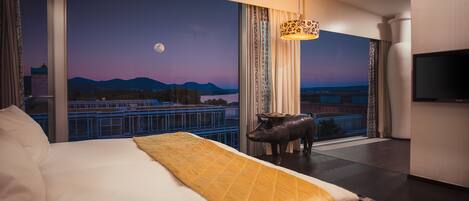 A hotel room with a large bed, a view of the cityscape at dusk, a mounted pig sculpture, and a television on the wall.