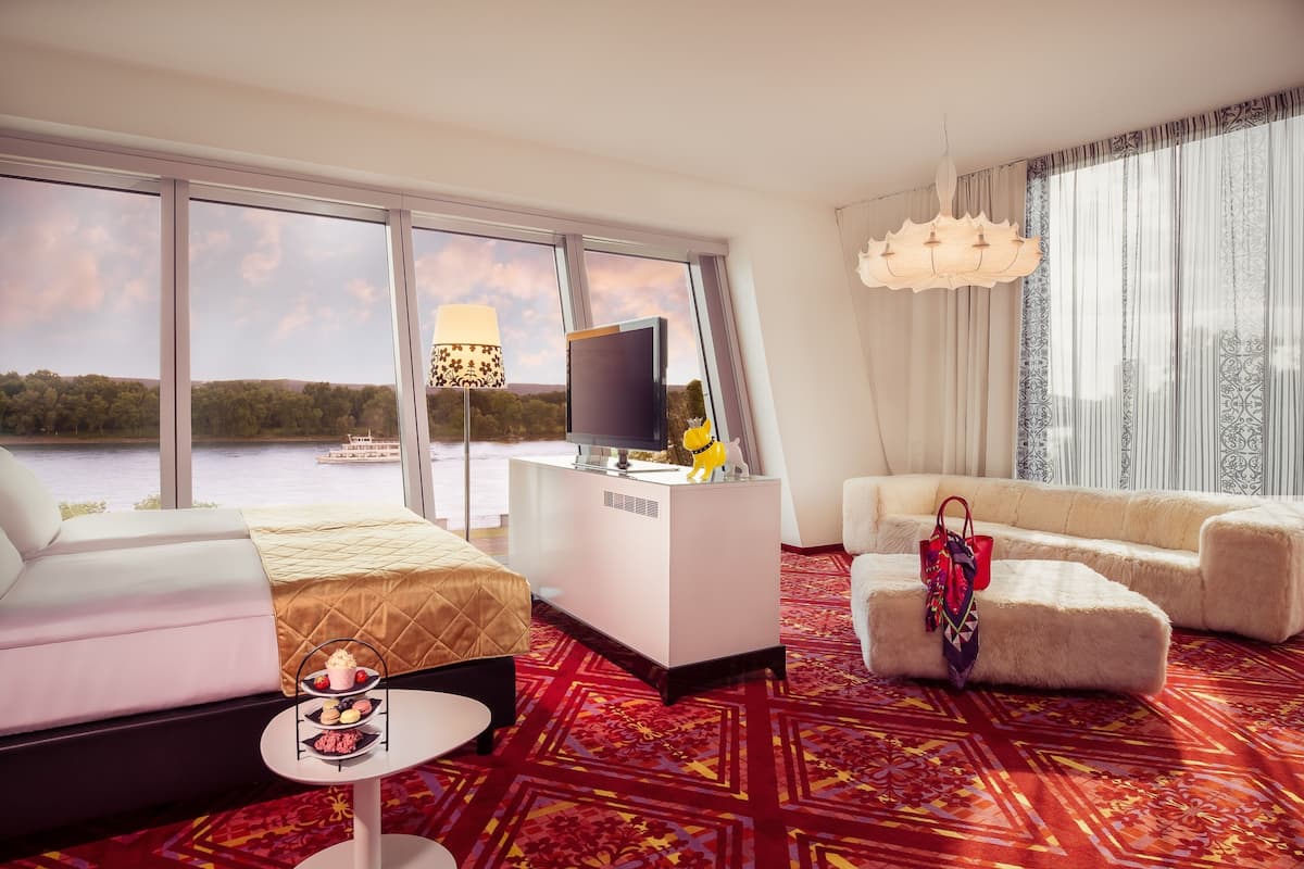 A hotel room with a bed, a sofa, a desk with a TV, a lamp, and a view of a lake.