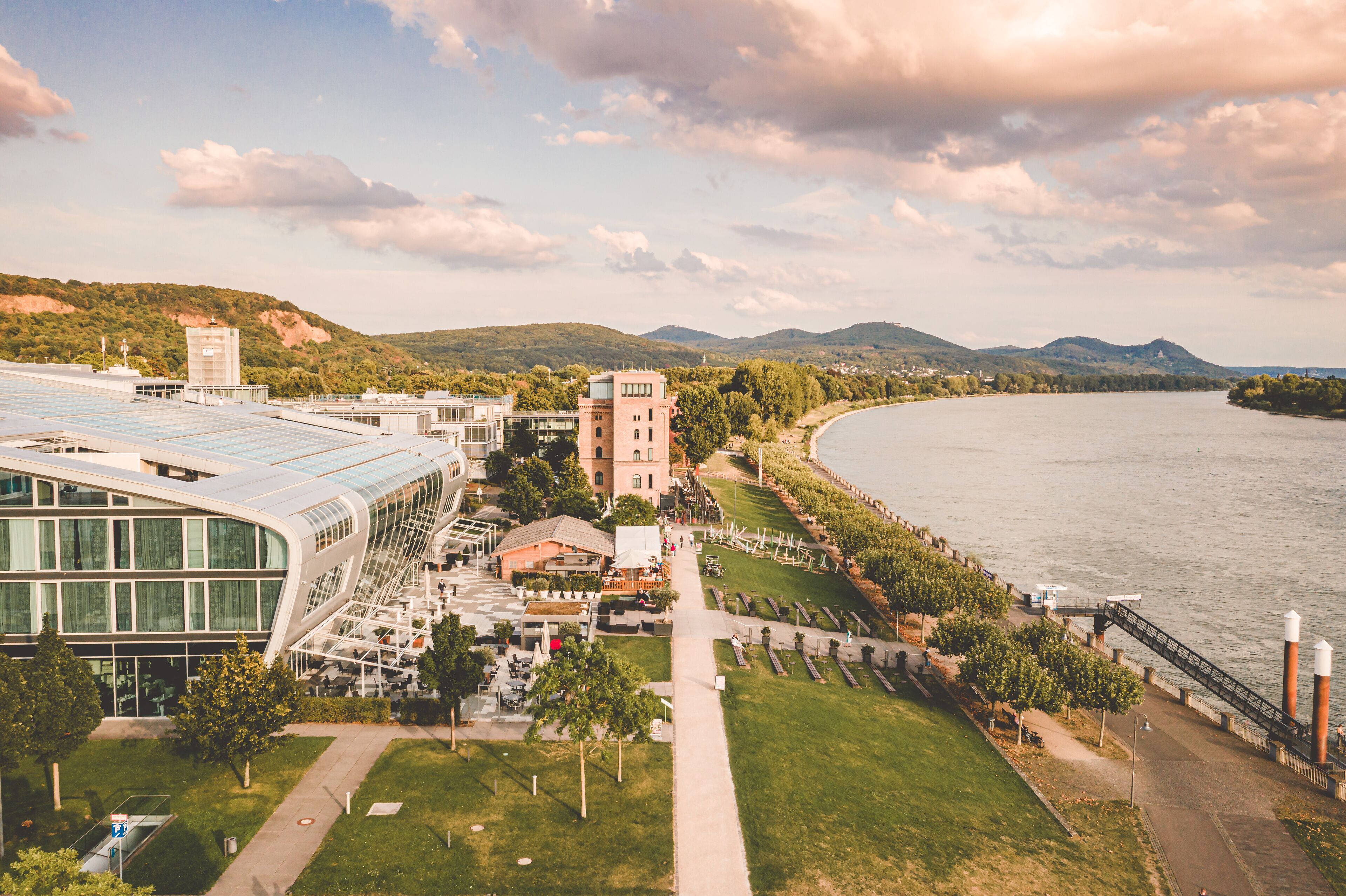 A riverside view of a modern building complex with a glass facade, a central tower, and a pedestrian walkway along the river.