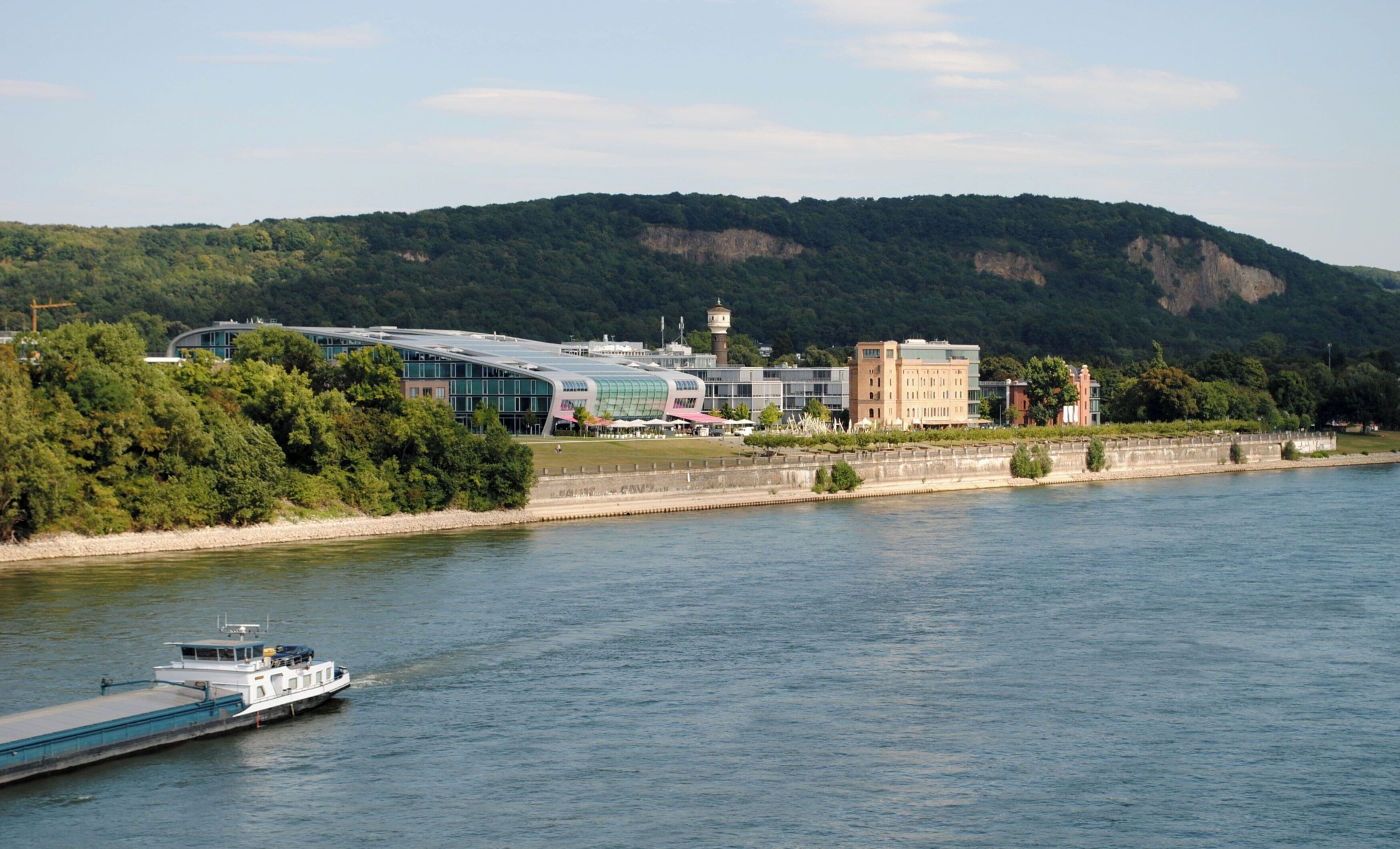A river with a boat, a modern building with a glass facade, and a forested hill in the background.