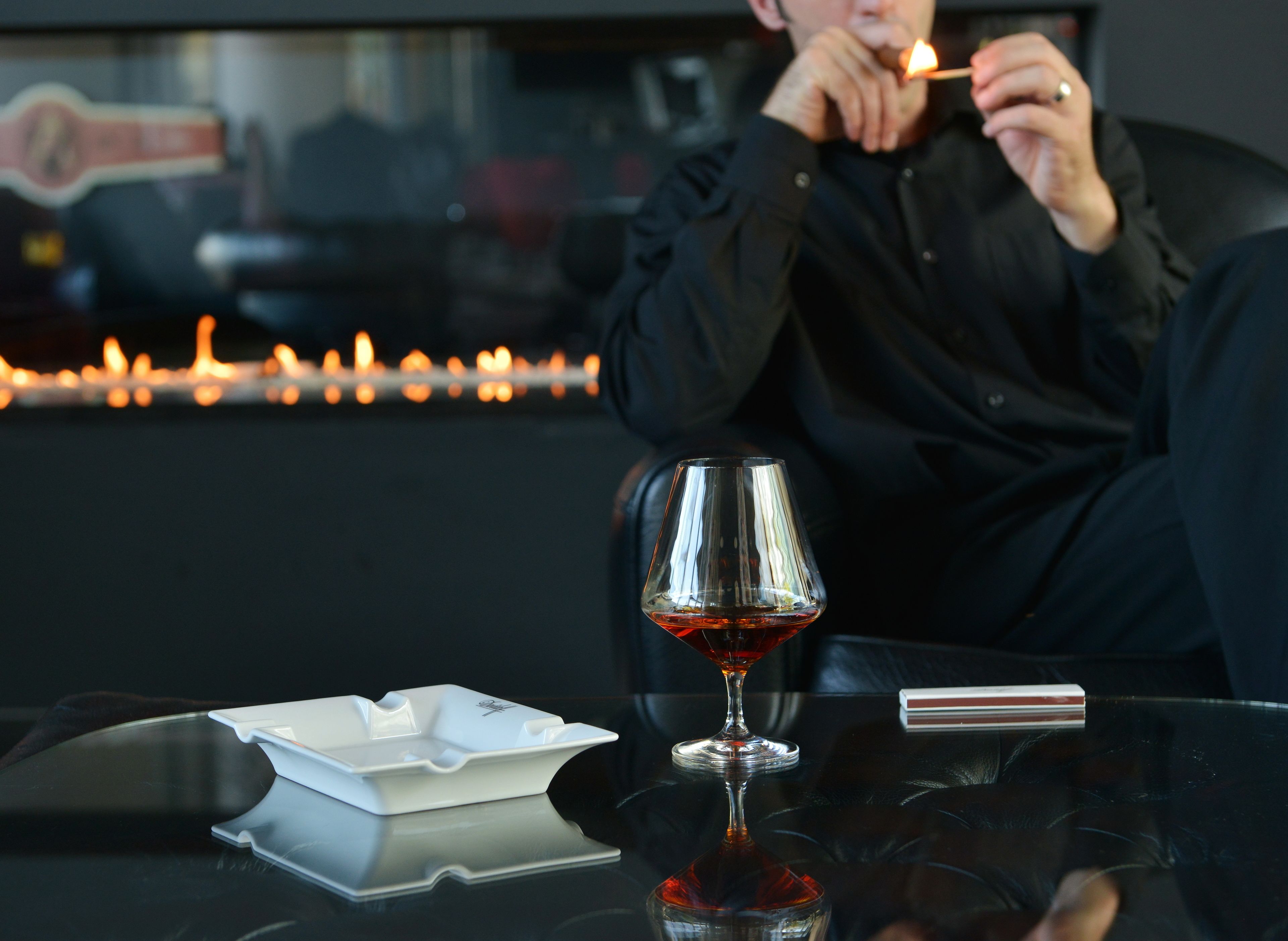 A person sitting at a table with a glass of red liquid, a white ashtray, and a lighter.
