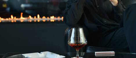 A person sitting at a table with a glass of red liquid, a white ashtray, and a lighter.