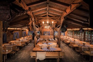 A rustic wooden interior with high ceilings, wooden benches, tables with checkered tablecloths, and a chandelier.