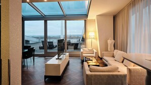 A modern hotel room with a large glass window, a sofa, a coffee table, and a lamp. - Kameha Grand Bonn (Bonn)