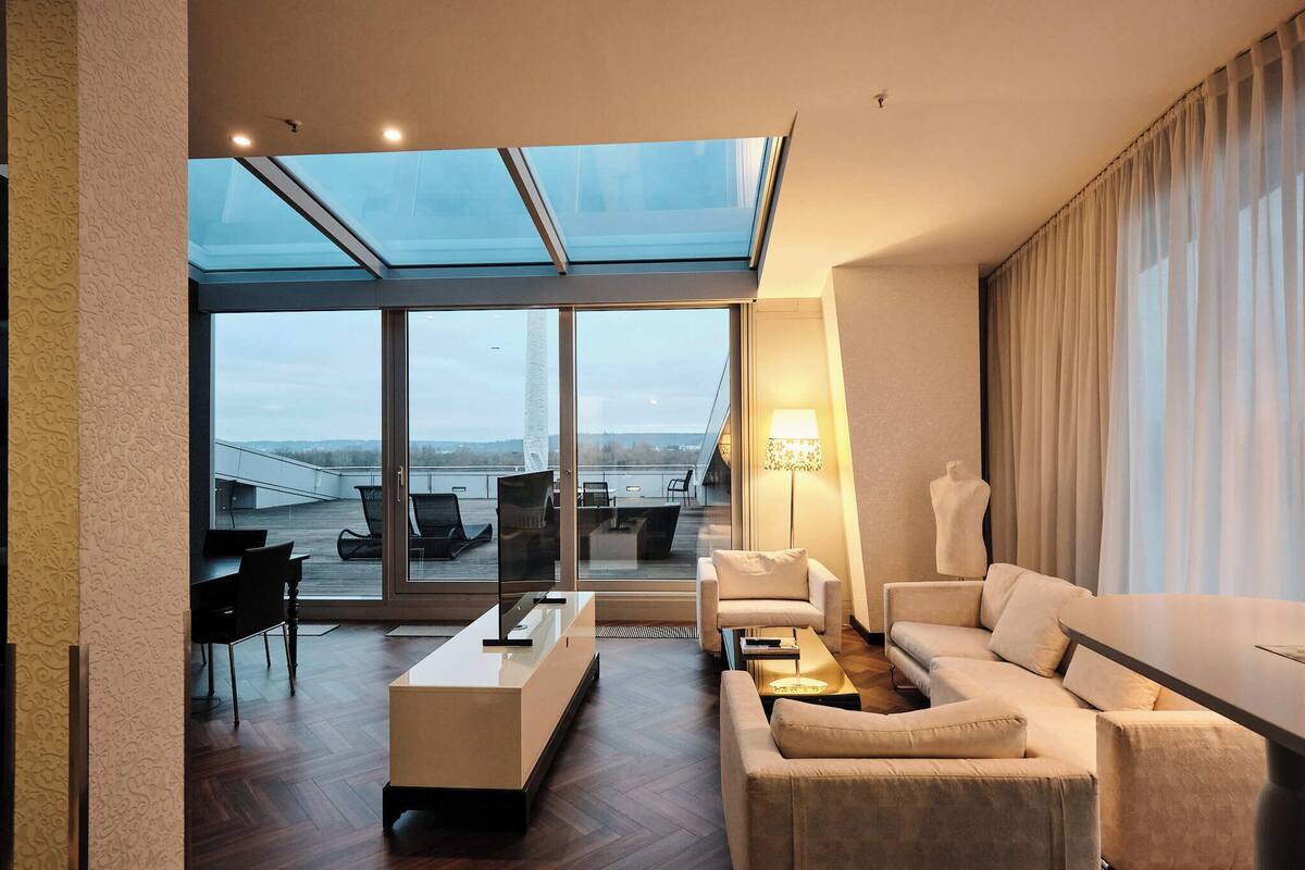 A modern hotel room with a large glass window, a sofa, a coffee table, and a lamp.