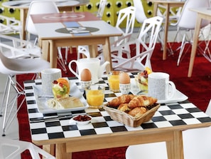 Une table de petit-déjeuner avec une nappe à carreaux, une théière, une tasse, une assiette de croissants, un panier de fruits et un panier de viennoiseries.