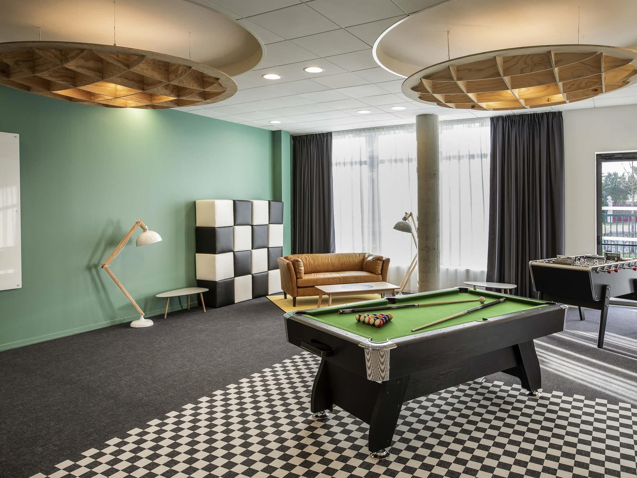 A pool table with a checkered floor, a sofa, a lamp, and a foosball table in a room with green walls and large windows.. ibis Styles Le Mans Sud Mulsanne