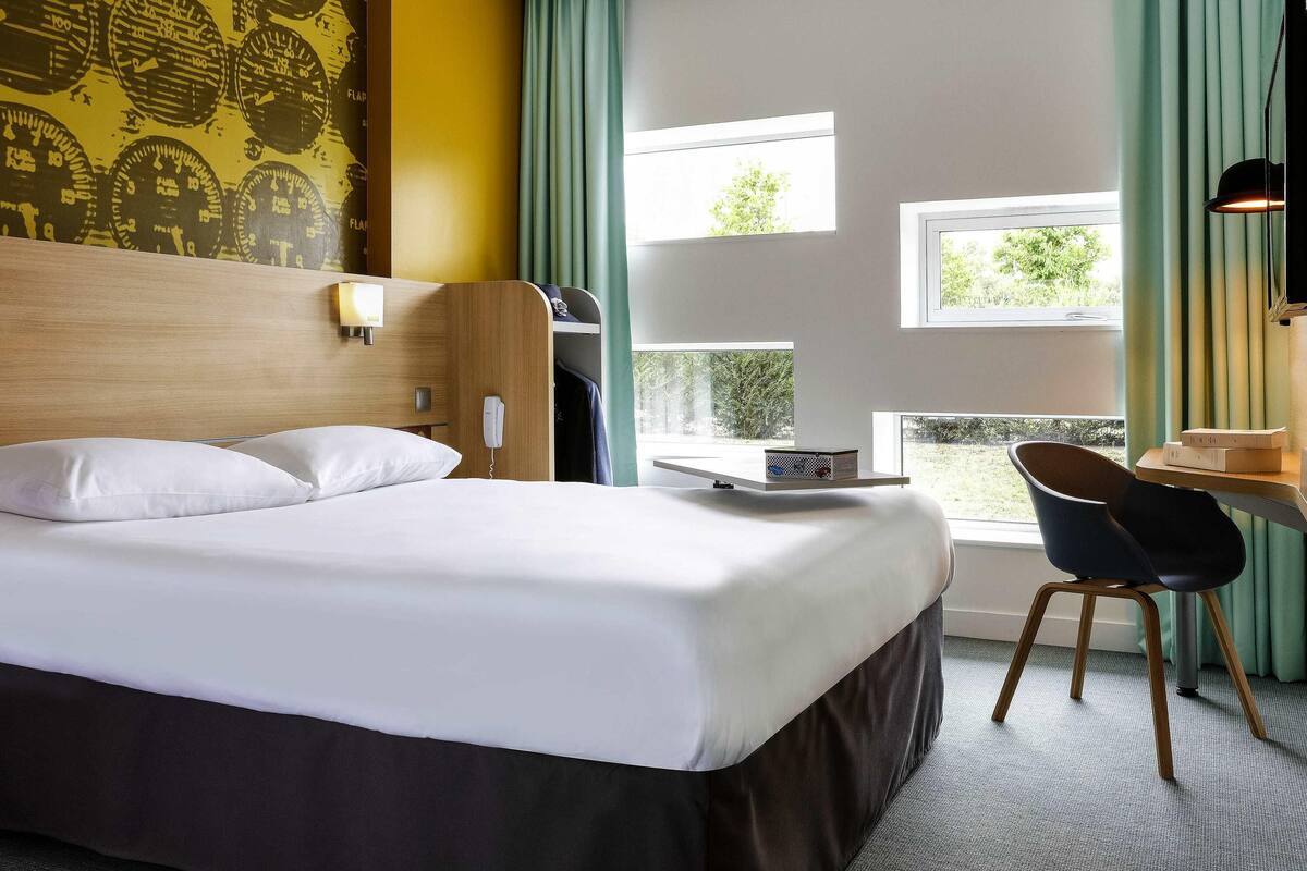 Een hotelkamer met een bed, een bureau met stoel, een telefoon en een raam met gordijnen.