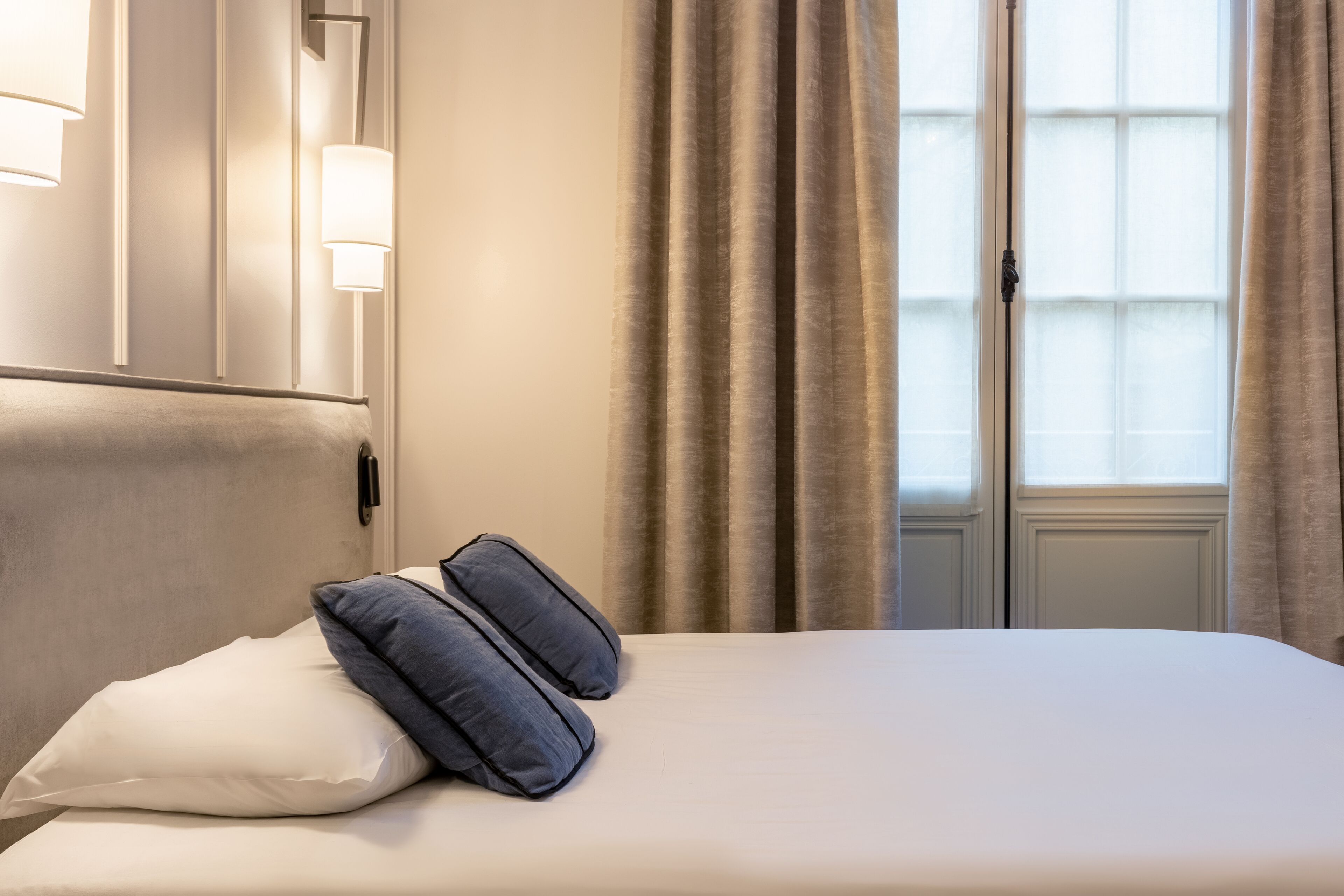 A hotel room with a bed, pillows, a nightstand, a lamp, and a window with curtains.