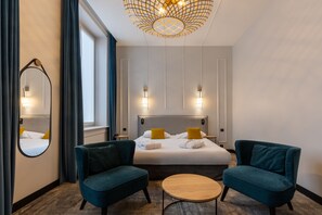 A hotel room with a large bed, two armchairs, a round coffee table, a mirror, and a chandelier.