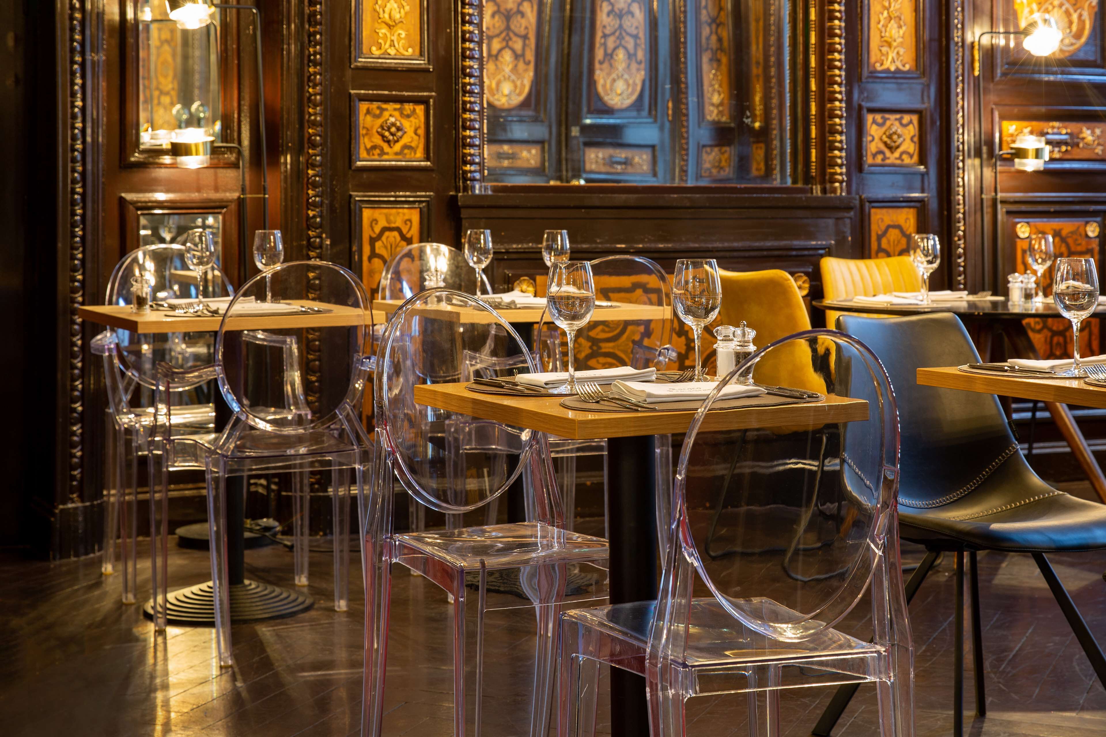 A restaurant interior with clear glass chairs, wooden tables, and ornate wall decorations.
