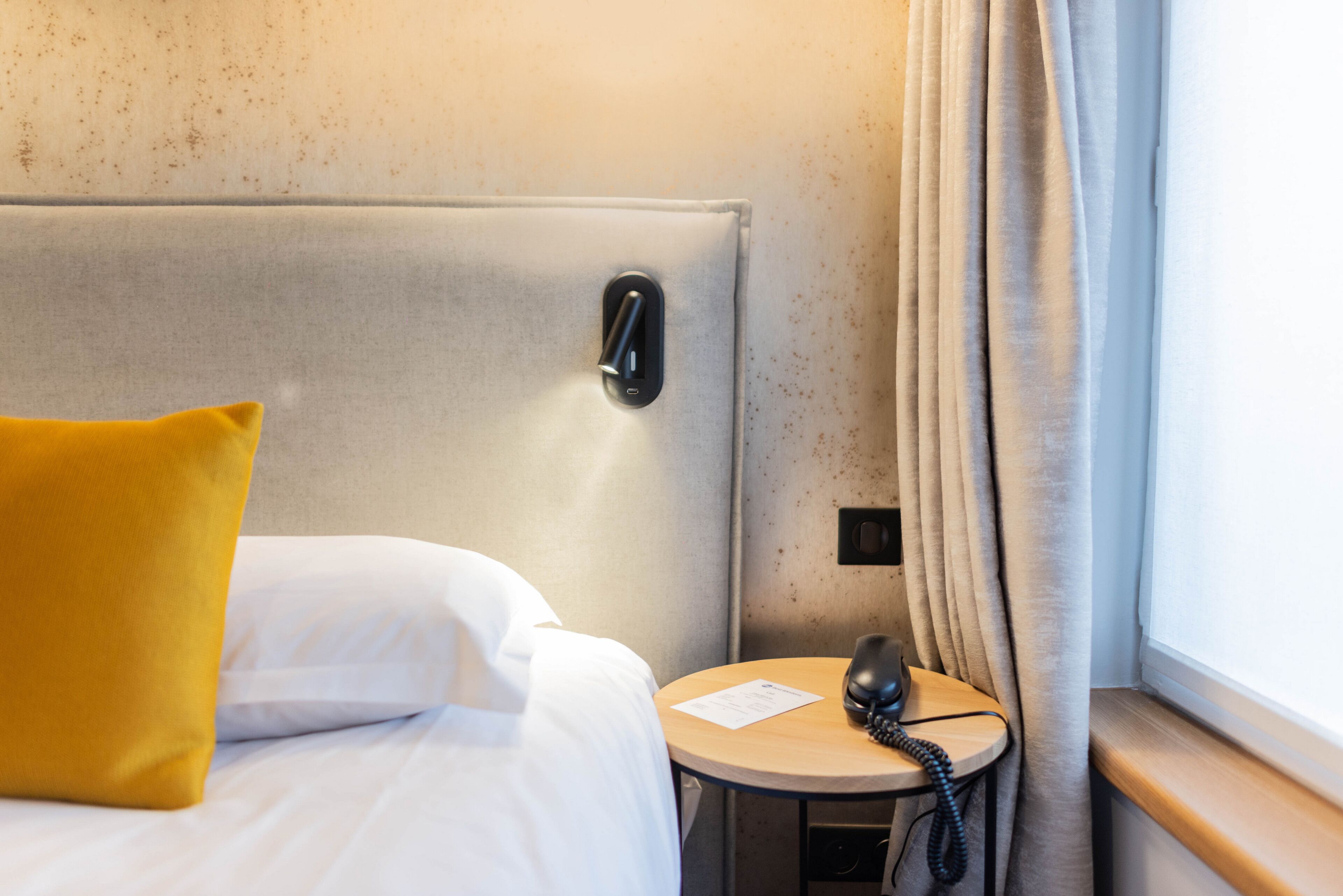 Une chambre d’hôtel avec un lit, un téléphone sur la table de chevet, une petite table ronde avec une lampe et une fenêtre avec des rideaux.