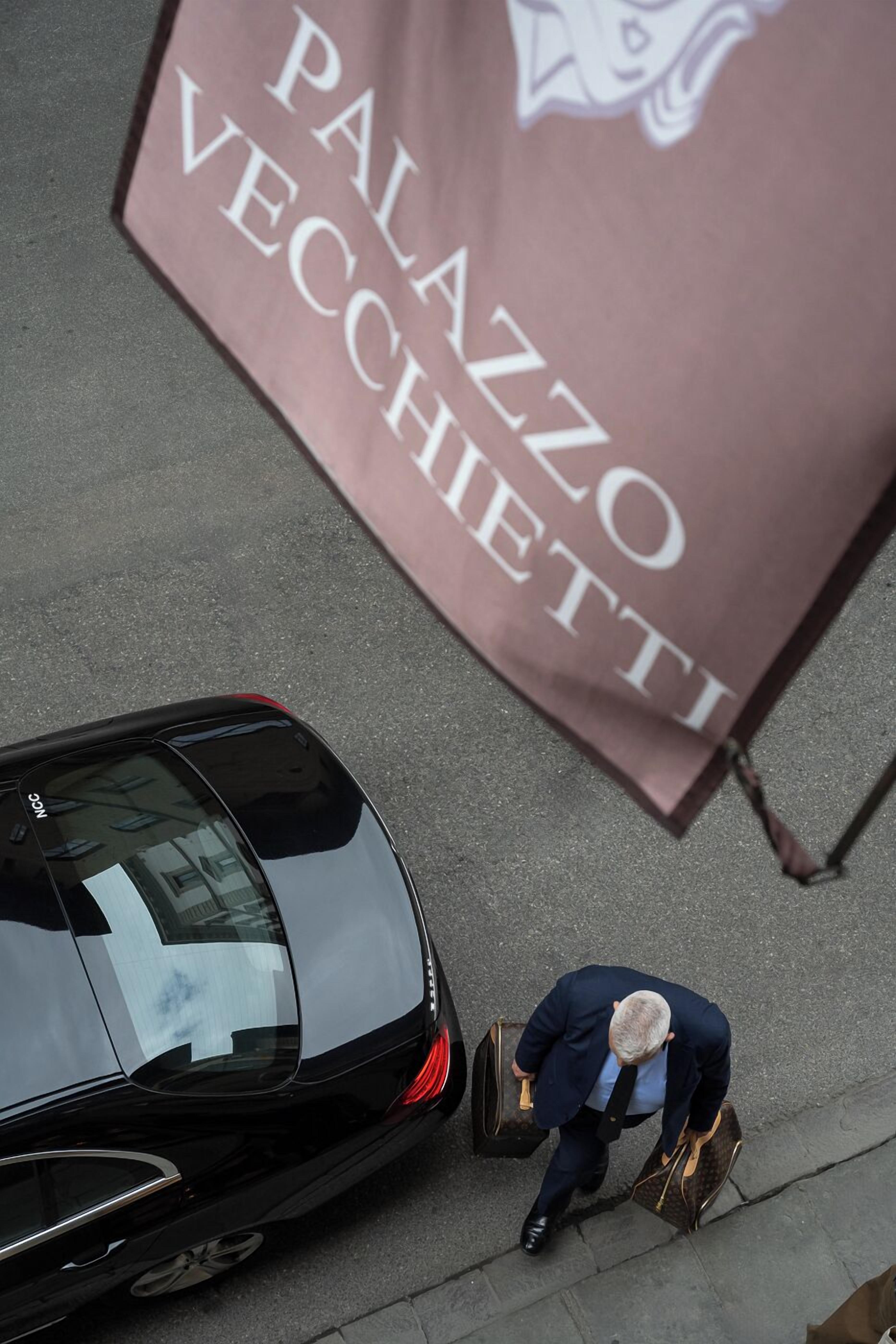 A man in a suit carrying a briefcase, walking past a black car and a flag with "PALAZZO VECCHETTI" text.