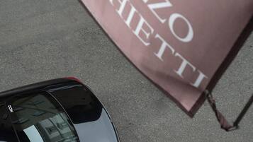 A man in a suit carrying a briefcase, walking past a black car and a flag with "PALAZZO VECCHETTI" text.