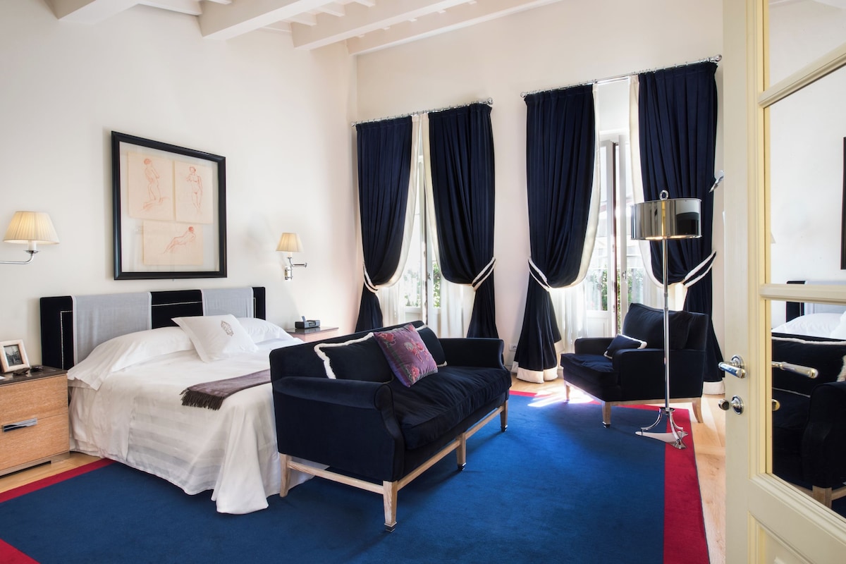 A hotel room with a bed, a black sofa, a chair, and a desk with a lamp.