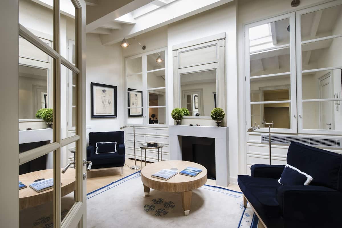 A spacious living room with a fireplace, a round coffee table with books, a dark blue sofa, and a clear view of the adjacent bedroom through a glass door.