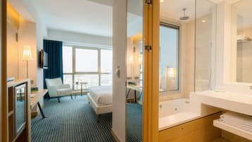 A hotel room with a bed, a desk, a chair, a large window, a bathroom with a bathtub, and a mirror.