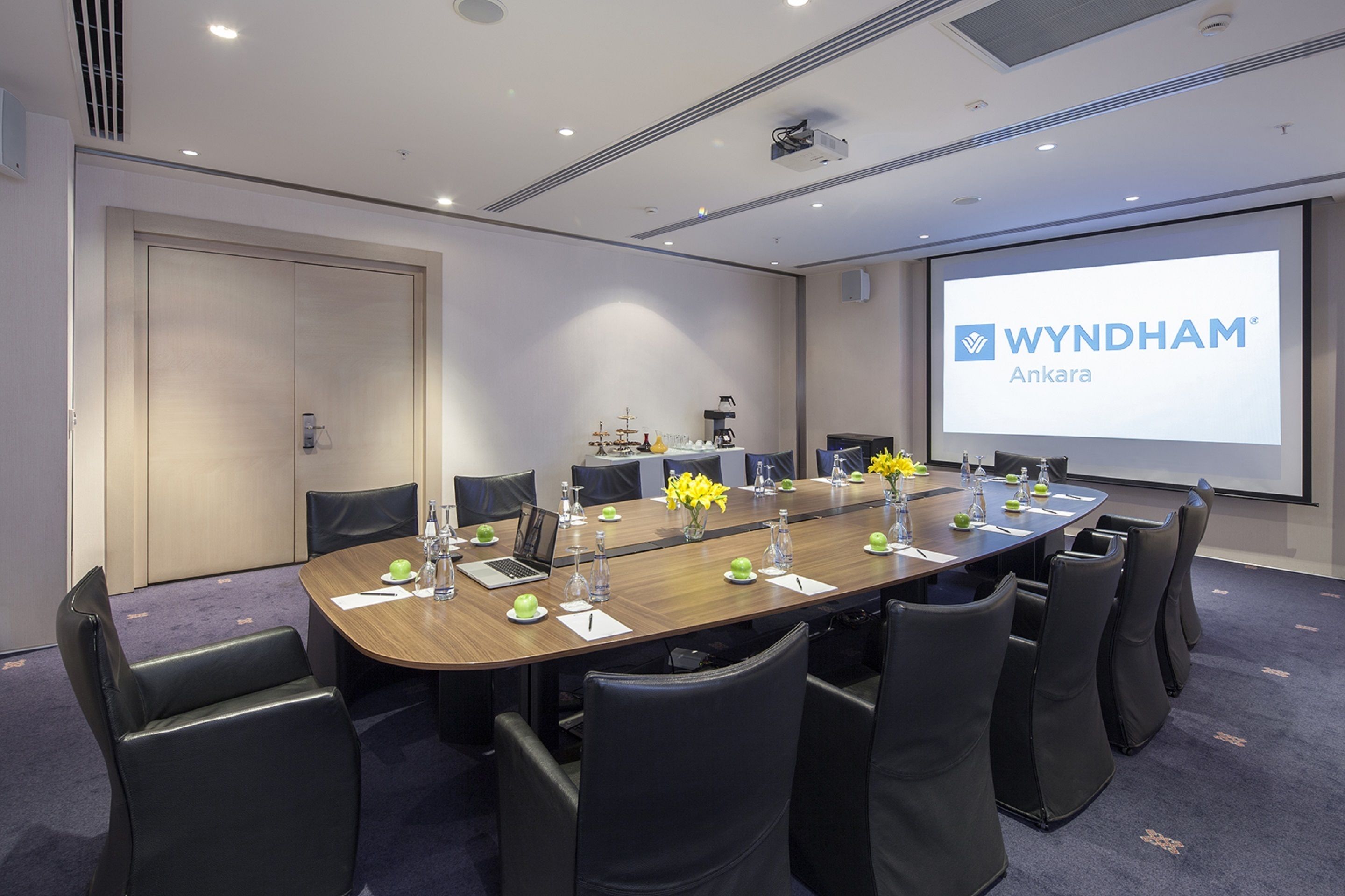 Un salón de conferencias con una mesa larga, sillas, una pantalla de proyector mostrando 'WYNDHAM Ankara' y una puerta cerrada.