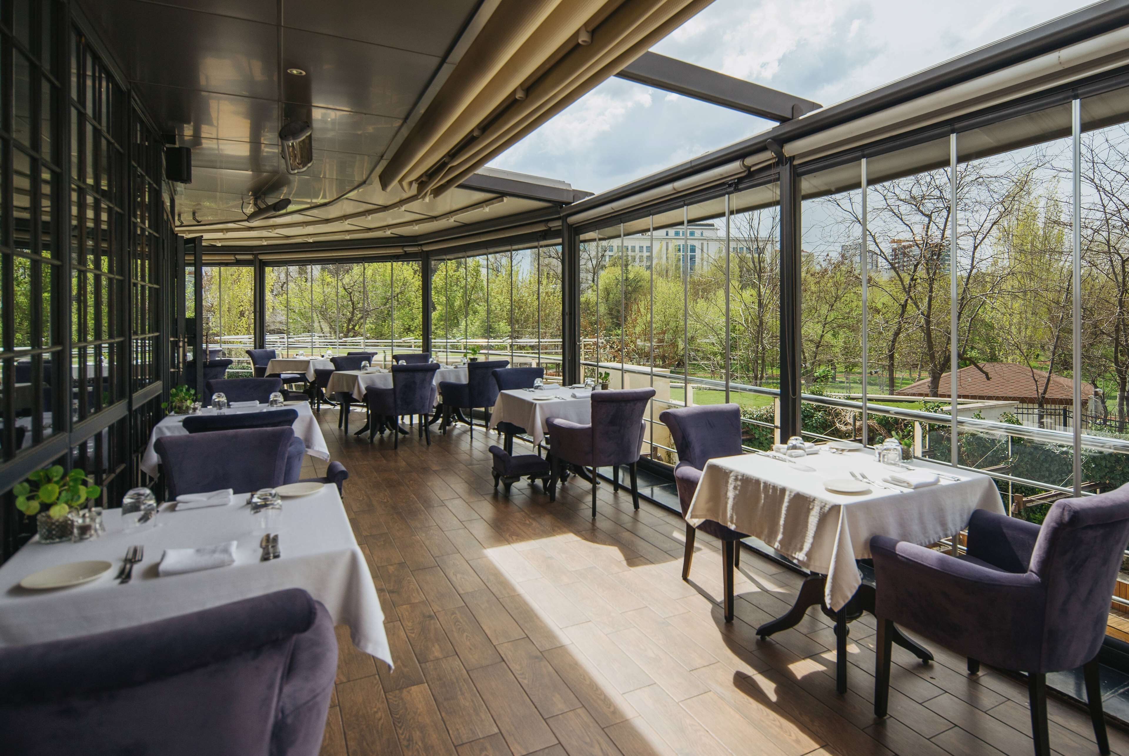 Un restaurante con mesas y sillas dispuestas en una terraza de madera, grandes ventanales que ofrecen vistas a árboles y edificios.