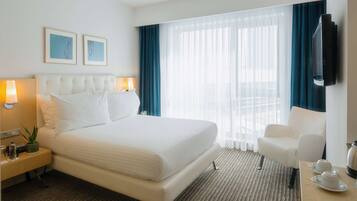 Quarto de hotel com uma cama grande, mesa de cabeceira com luminária, uma cadeira e televisão fixada na parede.