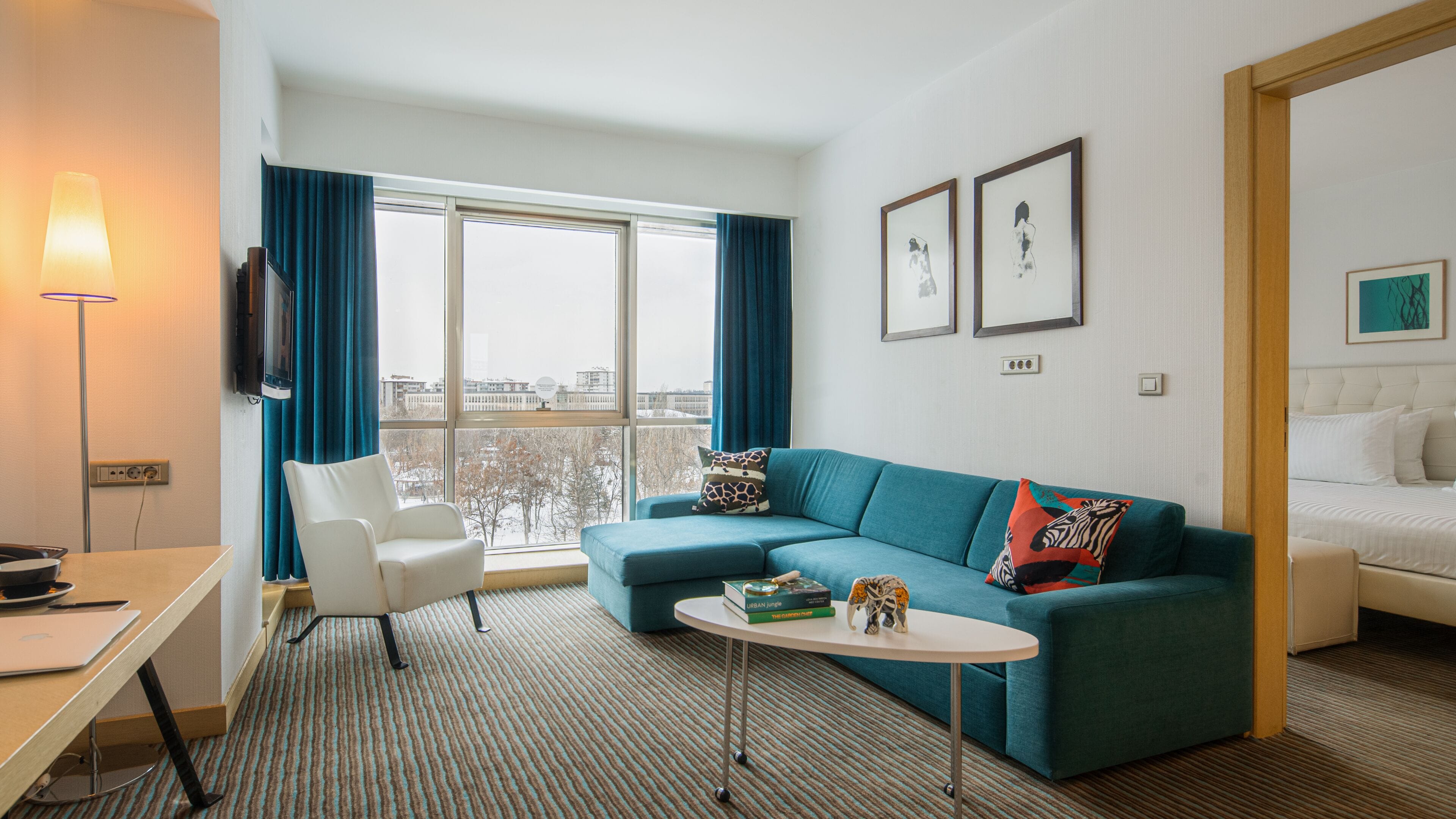 Quarto de hotel moderno com um sofá azul-esverdeado, uma poltrona branca, uma pequena mesa redonda e uma cama com roupa de cama branca.