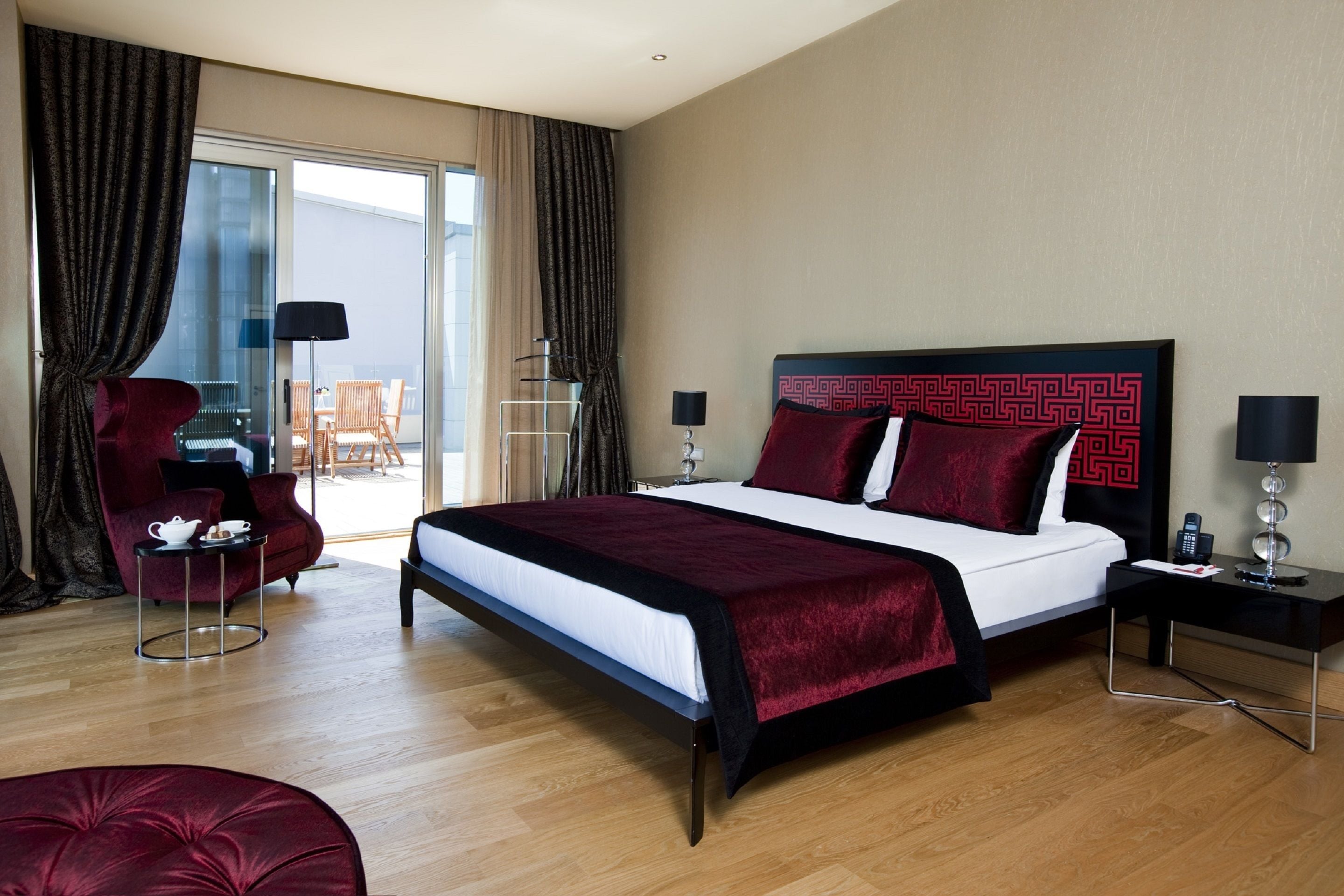 Una habitación de hotel moderna con una cama grande, una zona de estar con una silla roja, una mesita con una lámpara y vistas al exterior a través de una puerta de vidrio.