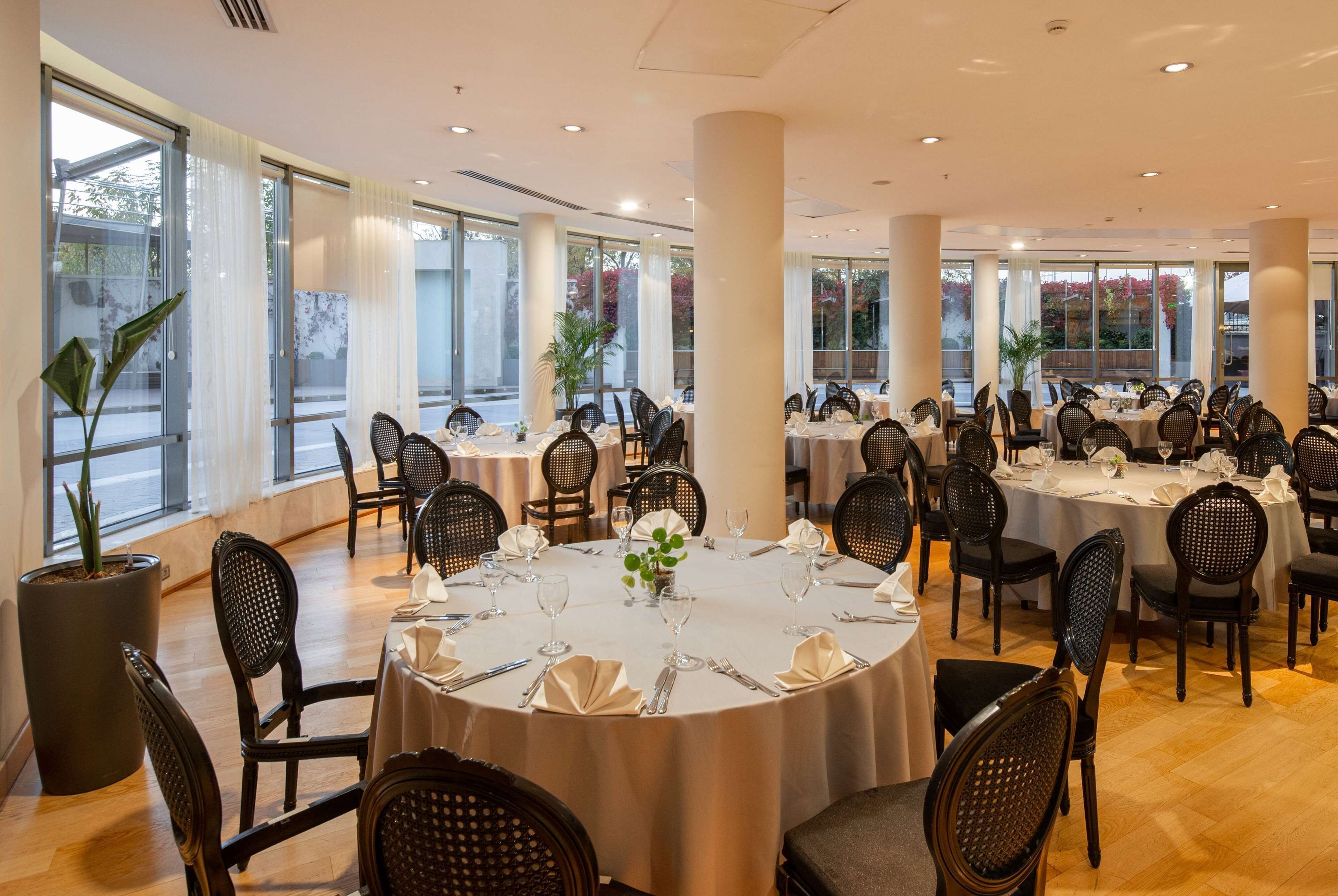 Un comedor amplio con mesas redondas, sillas de mimbre negro y ventanas grandes con vistas al exterior.