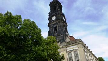Un edificio storico con una torre dell'orologio imponente, ampie finestre e alberi verdi rigogliosi in primo piano.