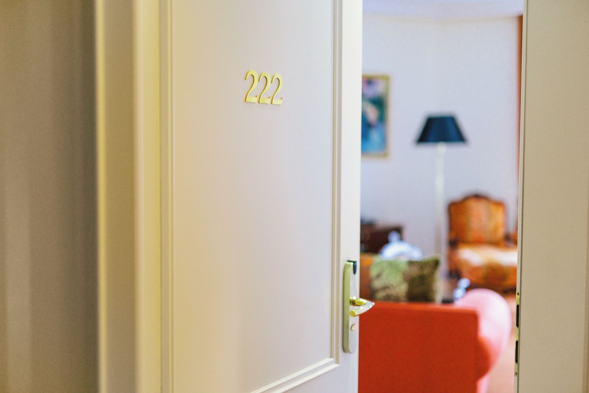 Eine Hotelzimmertür mit der Nummer 222, ein roter Sessel und eine Lampe im Hintergrund.