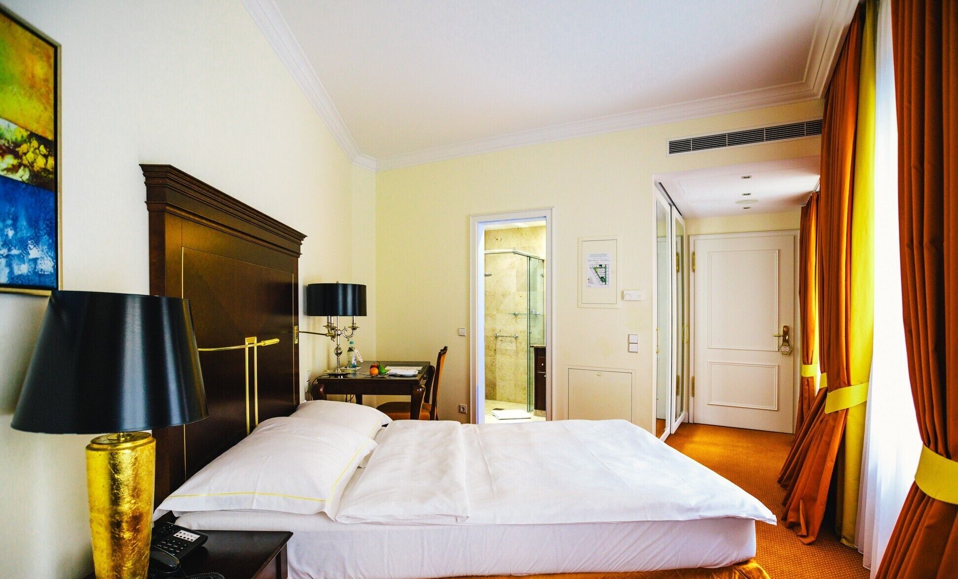 Ein Hotelzimmer mit einem Bett, einem Schreibtisch, einer Lampe, einem Gemälde und einer Tür zu einem weiteren Raum.