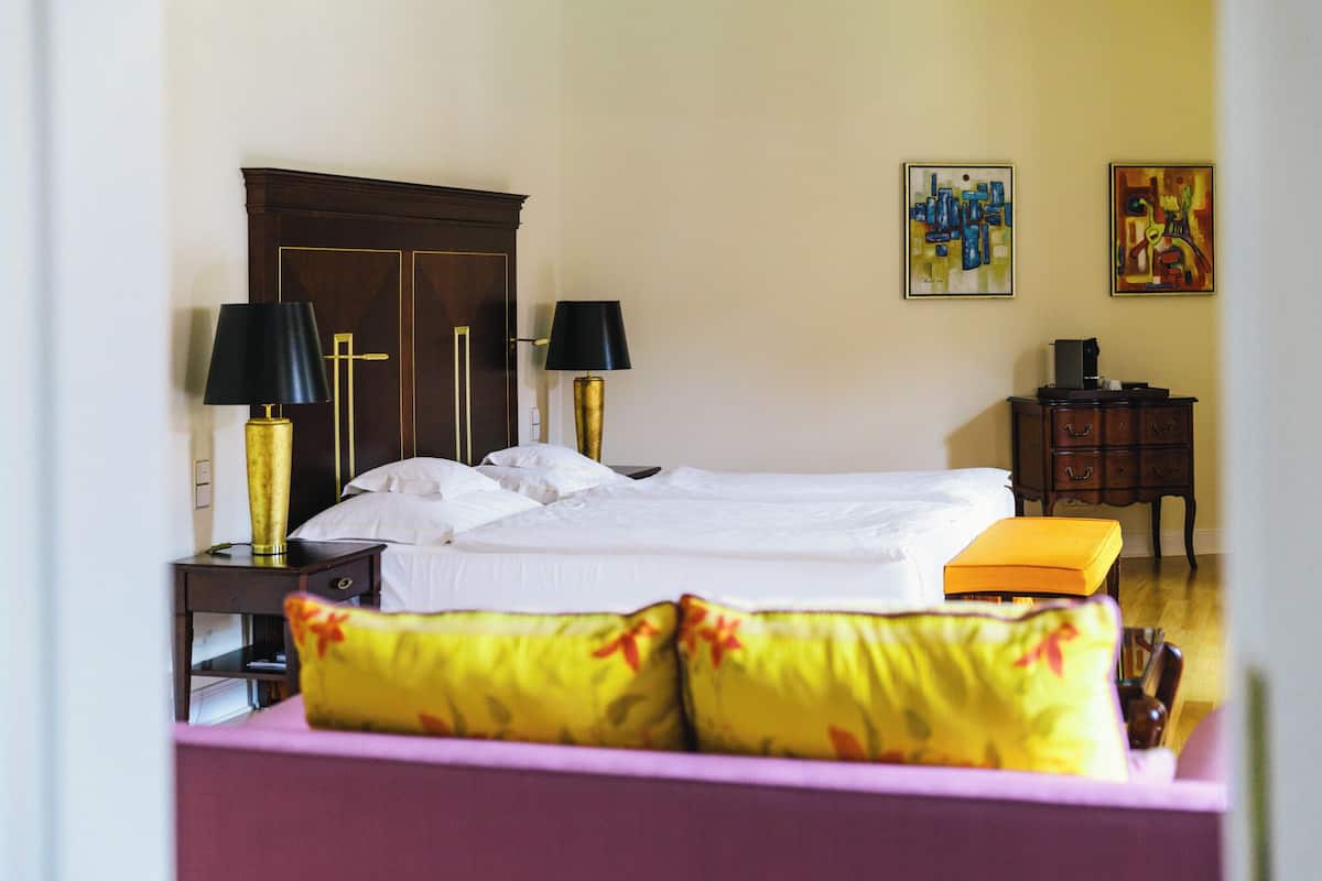 Habitación de hotel con cama, dos lámparas de noche, mesitas de noche, cómoda y dos cuadros en la pared.