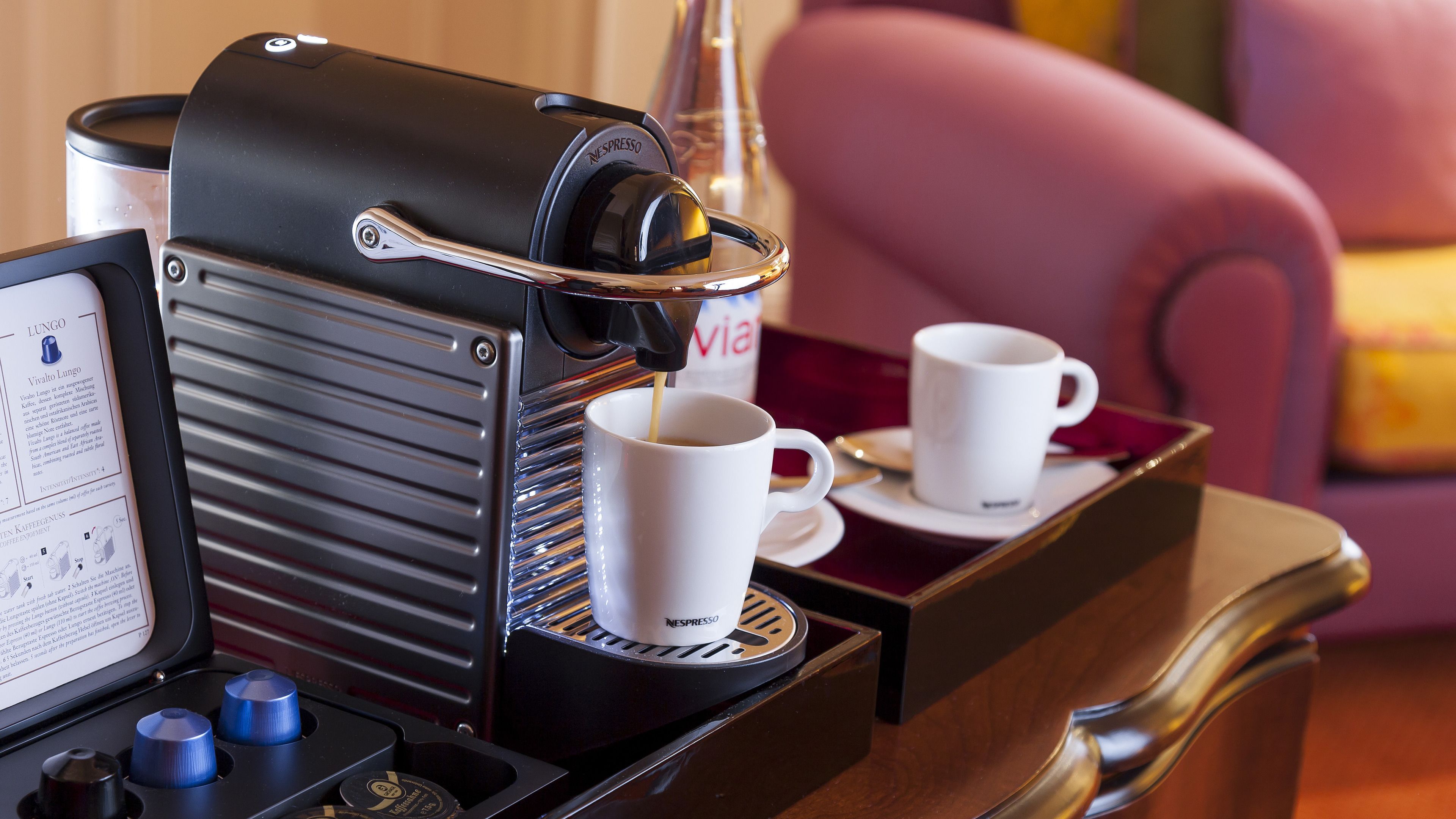 A coffee machine with two white cups on a tray, a menu, and a pink chair in the background.